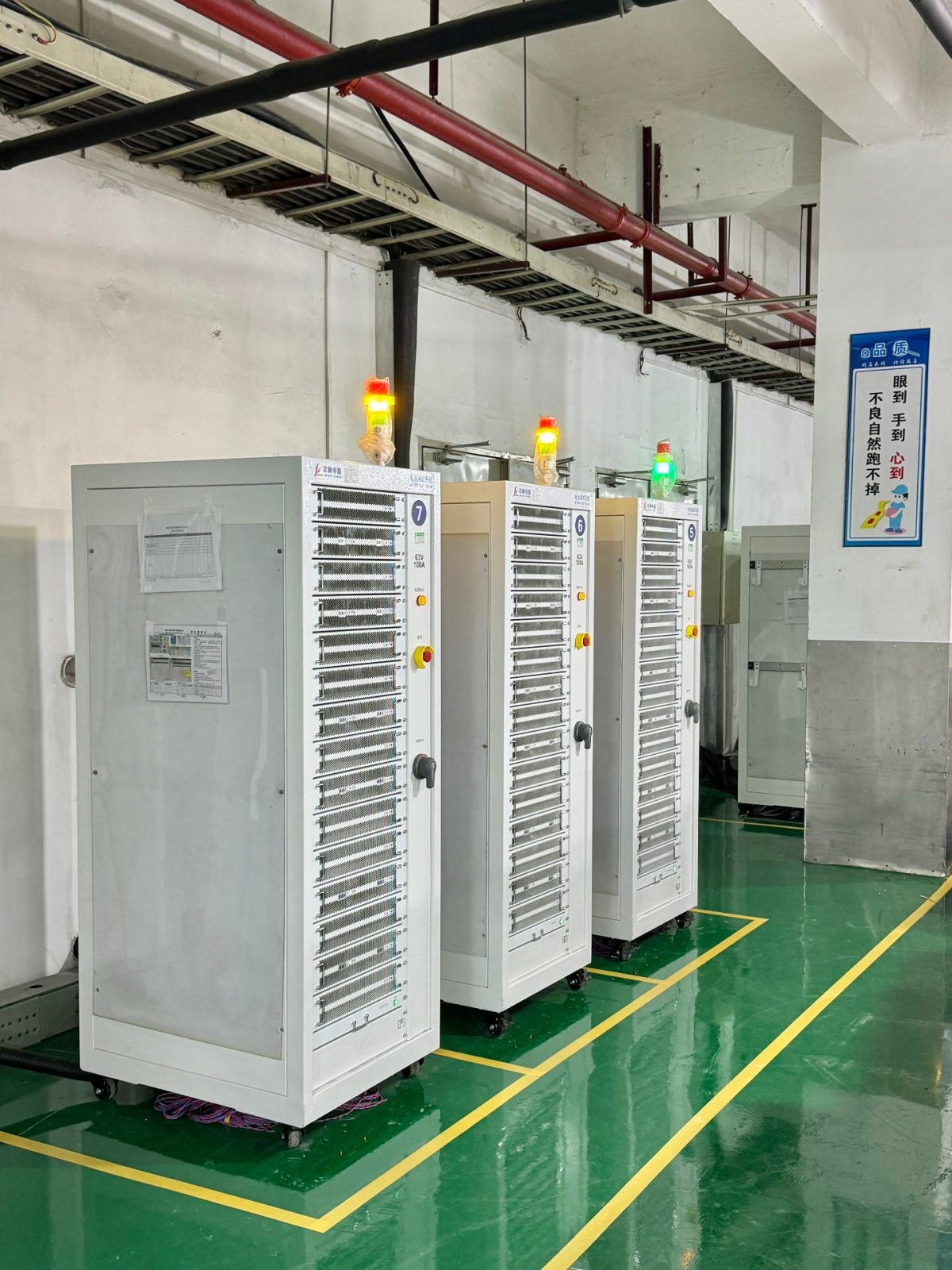 NovaBESS production line: row of formation and grading cabinets with yellow, red and green tower lights and floor safety markings